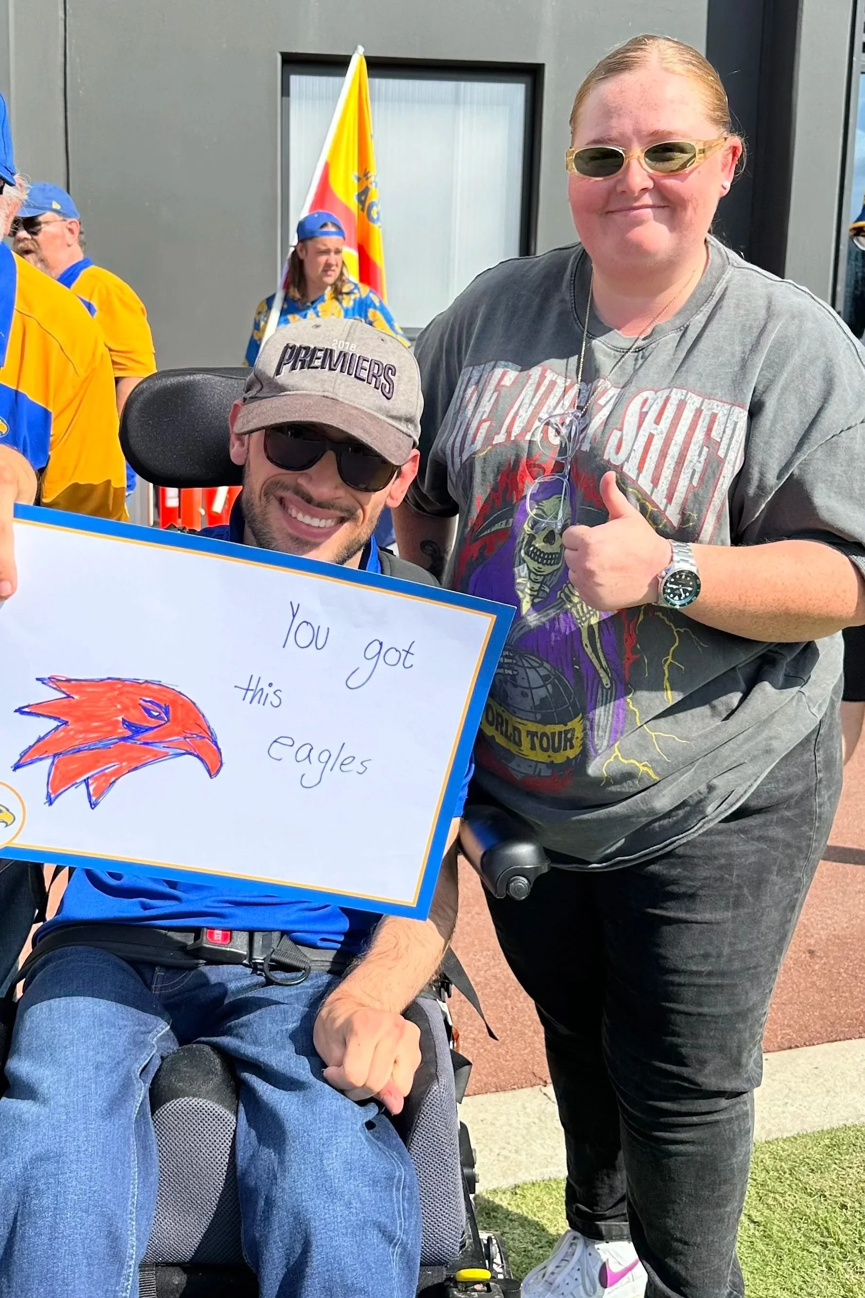 A man in a wheelchair holding a sign that says "You got this eagles" with a drawing of an eagle's head, smiling, wearing sunglasses, and two women standing behind him giving a thumbs-up at an outdoor event with fans wearing sports jerseys and flags i