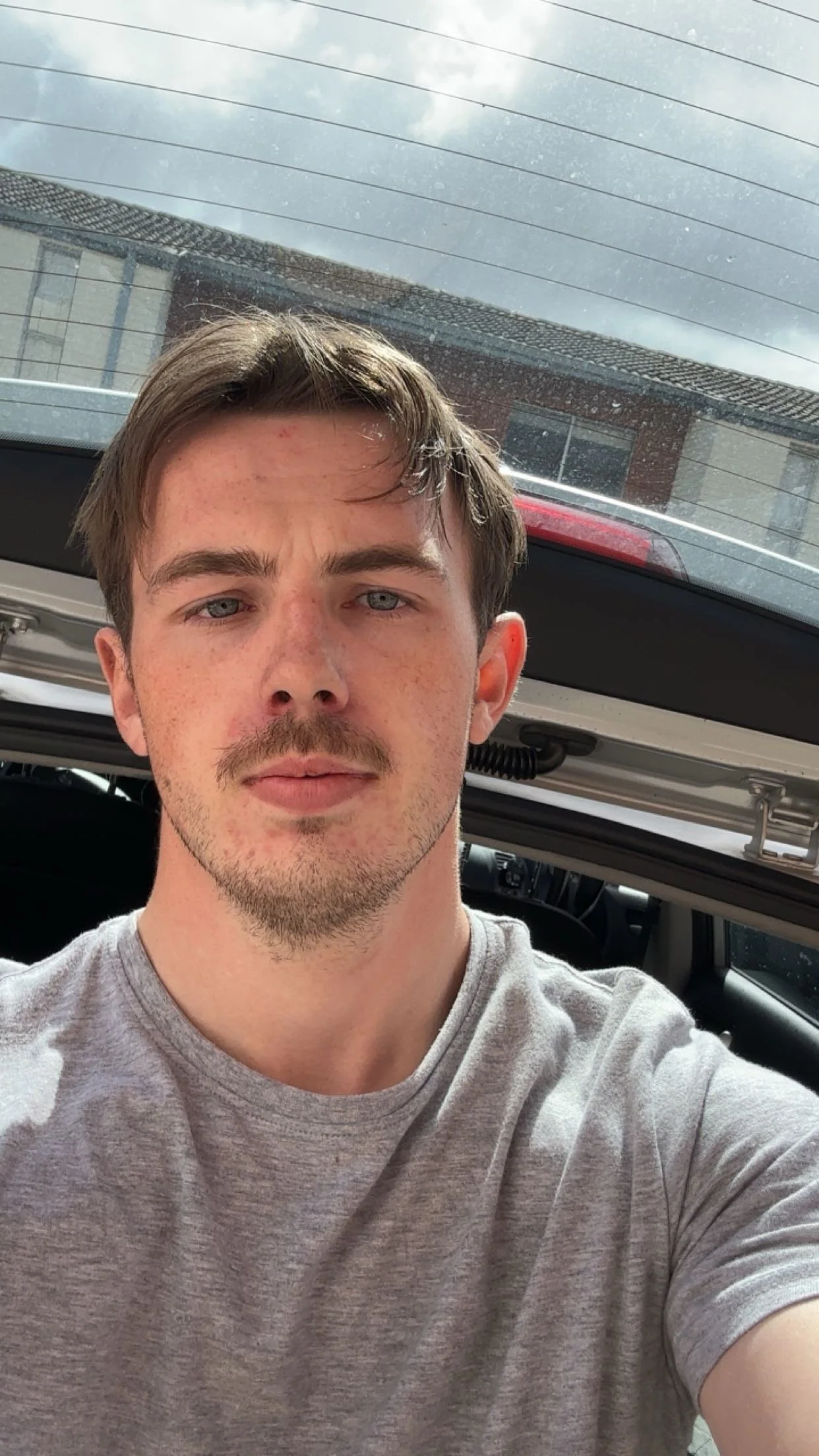 A young man with short brown hair, blue eyes, and light skin taking a selfie inside a vehicle with the back hatch open, showing buildings and a cloudy sky outside.