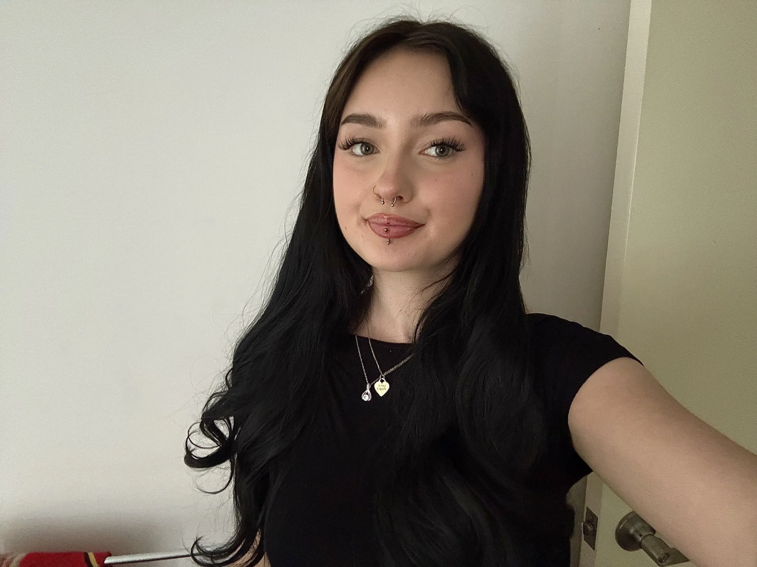 A young woman with long dark hair, wearing makeup and jewelry, taking a selfie in front of a plain beige wall.