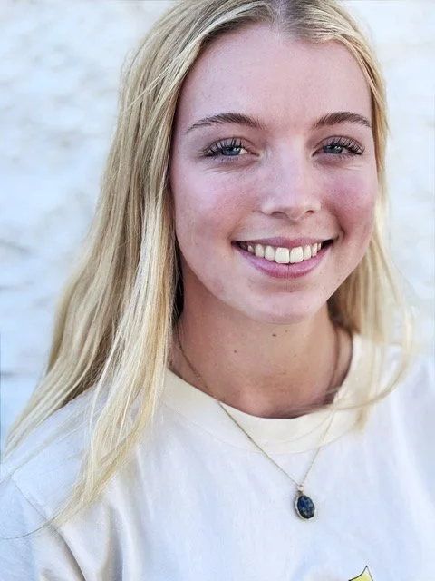Close-up photo of a young woman with long red hair, smiling, wearing a light blue and white sweatshirt, standing in front of a yellow brick wall.