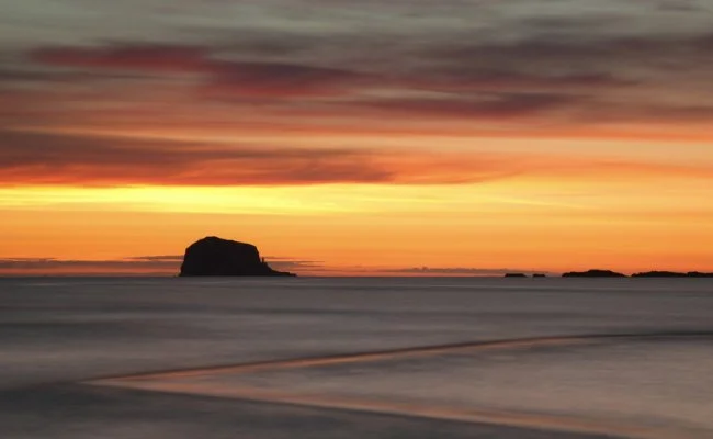 Sunset over the ocean with a silhouette of a large rock formation and smaller rocks in the distance, calm water, and a colorful sky with shades of orange, yellow, and pink.