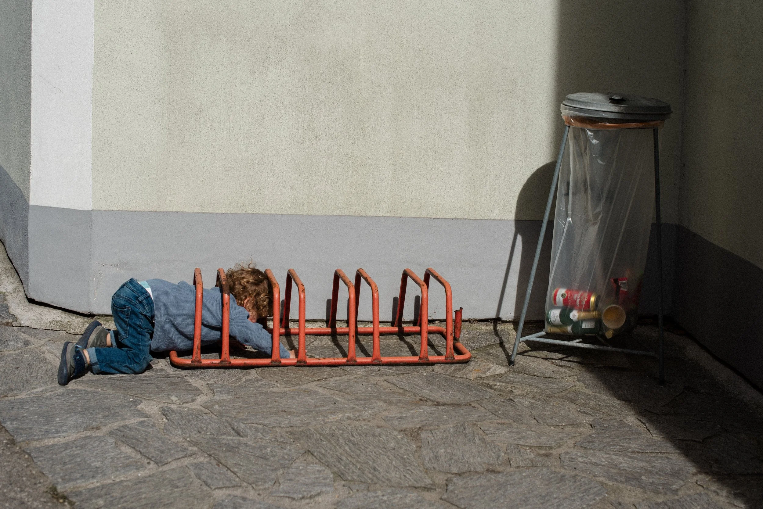 Ein Kind liegt auf dem Boden auf dem Bauch, spielt mit einem roten Fahrradständer, neben einer Mülltonne mit einer transparenten Abdeckung, in der alte Dosen und Verpackungen liegen.
