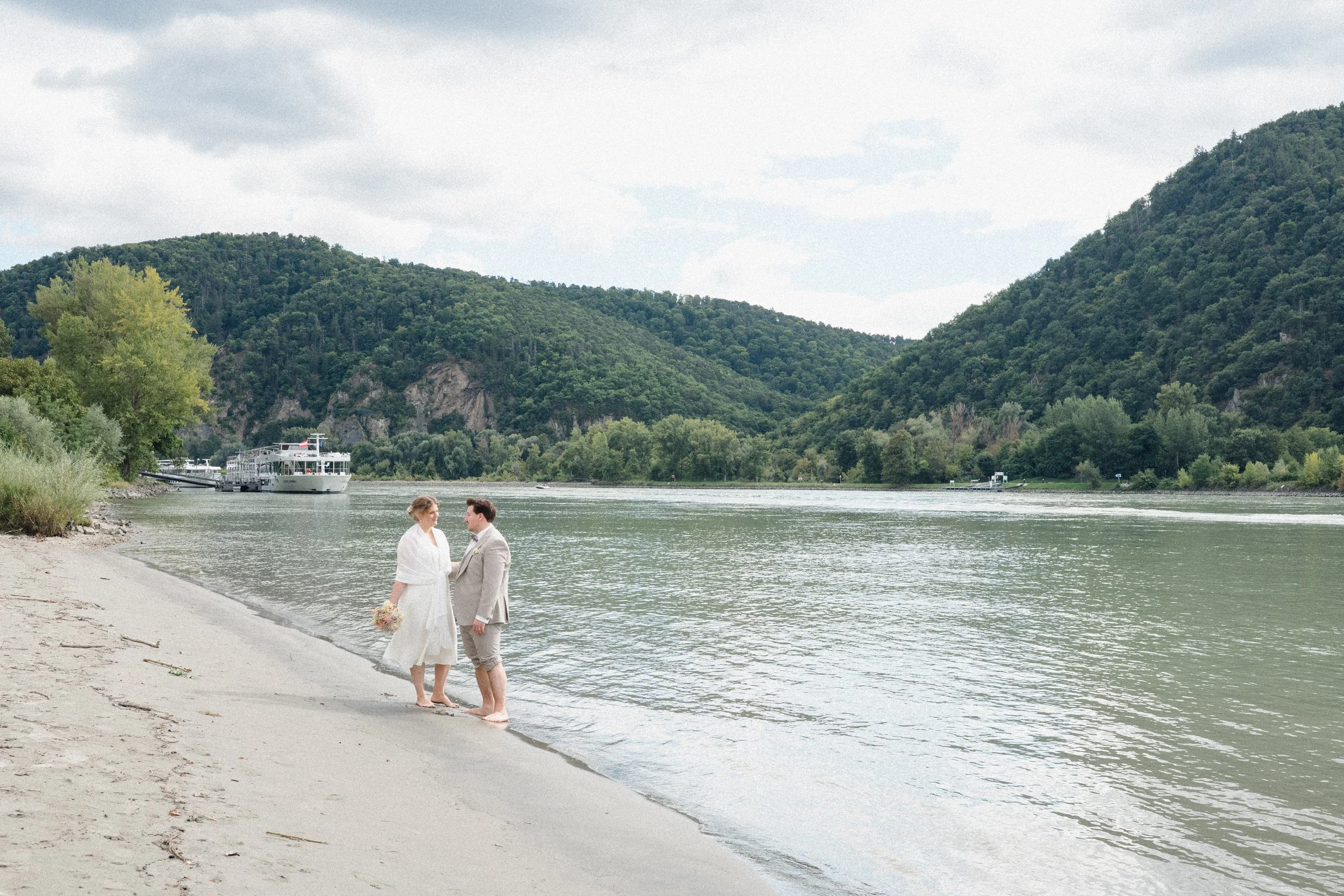 Ein heiratswilliges Paar steht am Flussufer, umgeben von grünen Hügeln, mit einem Boot im Hintergrund. Hochzeitsfotos nach standesamtlicher Trauung.