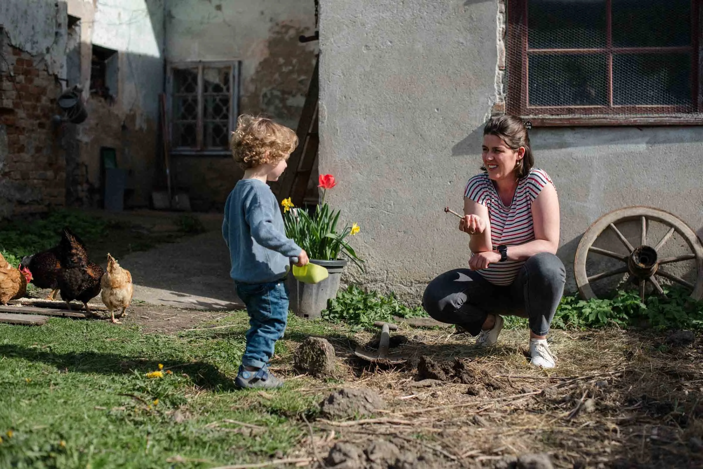 Eine Frau und ein Kind im Garten, die mit Hühnern und einer Blumentopf mit Blumen interagieren. Die Frau sitzt vor einer ungepflügten Erde, das Kind hält eine Gießkanne.
