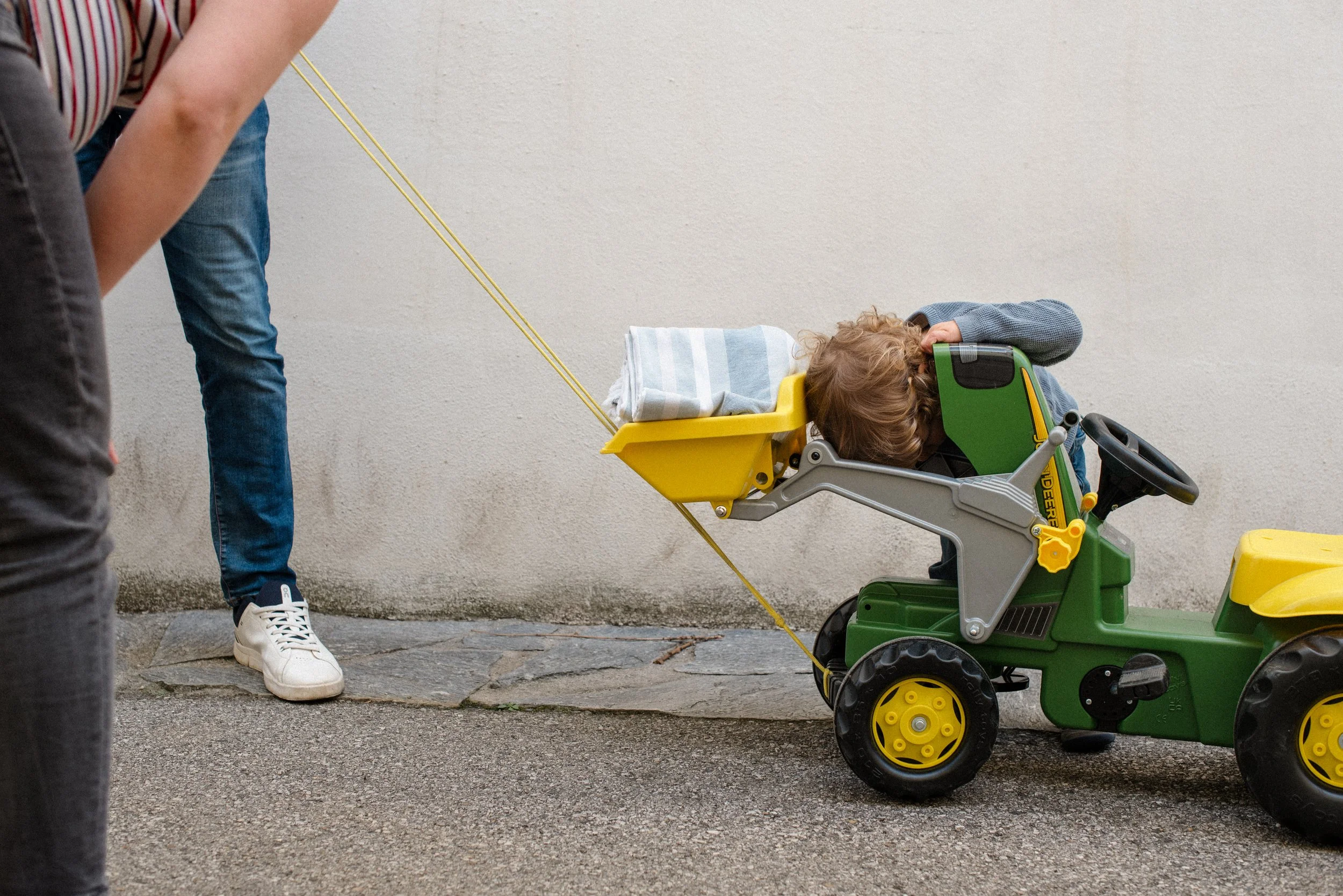 Ein Kind liegt auf einem kleinen Spielzeugtraktor, der an einer Leine gezogen wird, während eine Person im Hintergrund an der Leine zieht.