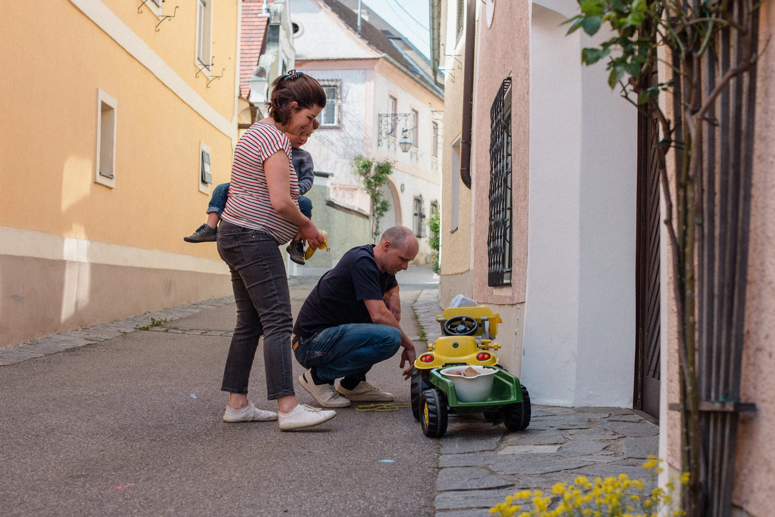 Familie spielt mit einem Kinderrasenmäher auf der Straße vor ihrem Haus in einer ruhigen Gasse.