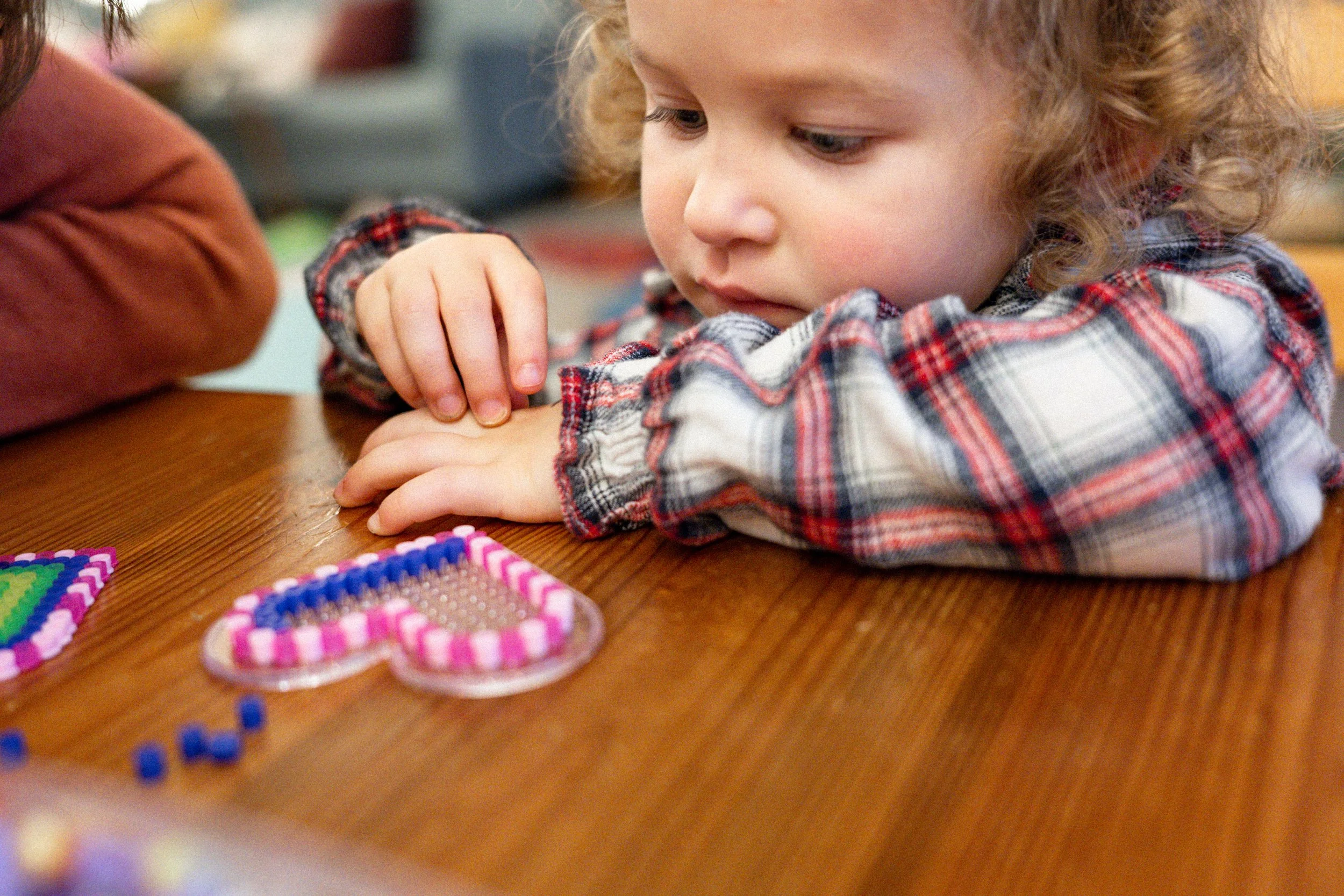 Ein Kind mit lockigem Haar und kariertem Hemd arbeitet mit Perlen und Perlenarmbändern am Tisch.
