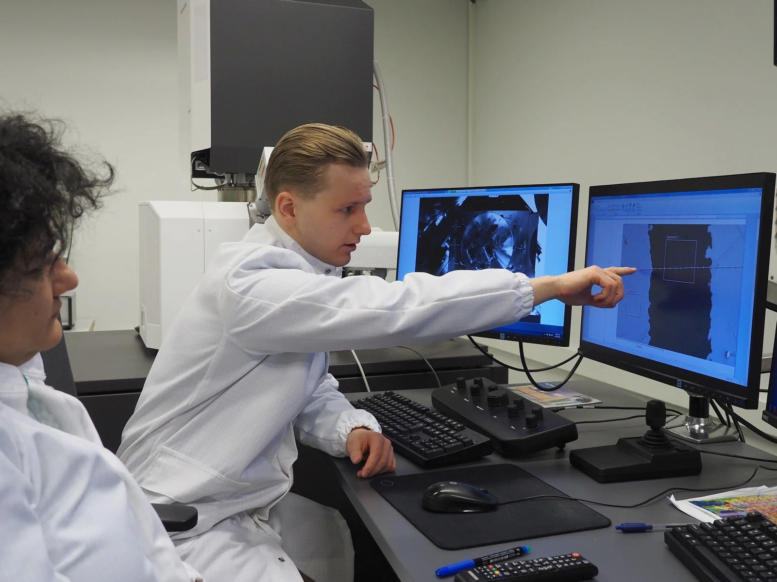Scientist in cleanroom suit analyzing semiconductor surface data on dual monitors in a high-tech lab