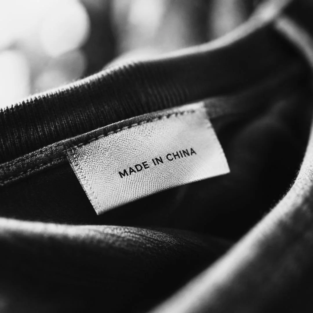 Black-and-white close-up of a clothing label reading “Made in China,” sewn inside a dark garment; soft focus and high contrast.