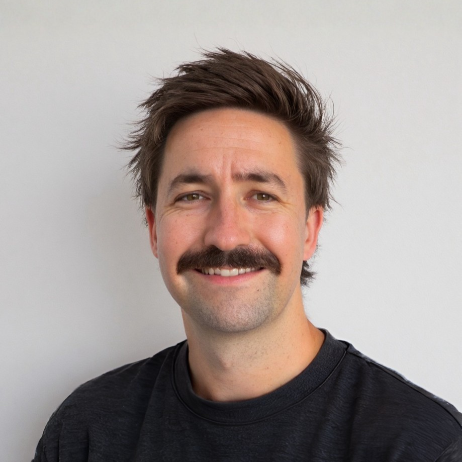 A man with brown hair, a mustache, and a beard wearing a black shirt, smiling against a plain white background.