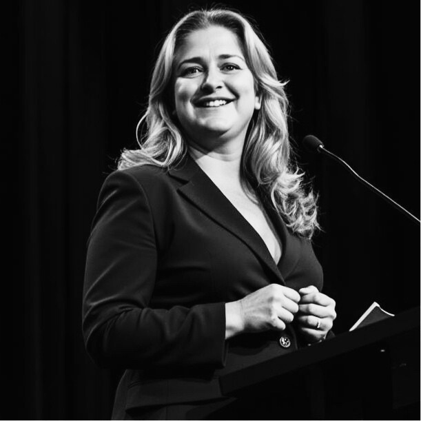 Smiling person in business attire speaking into a microphone at a podium, black and white photograph.