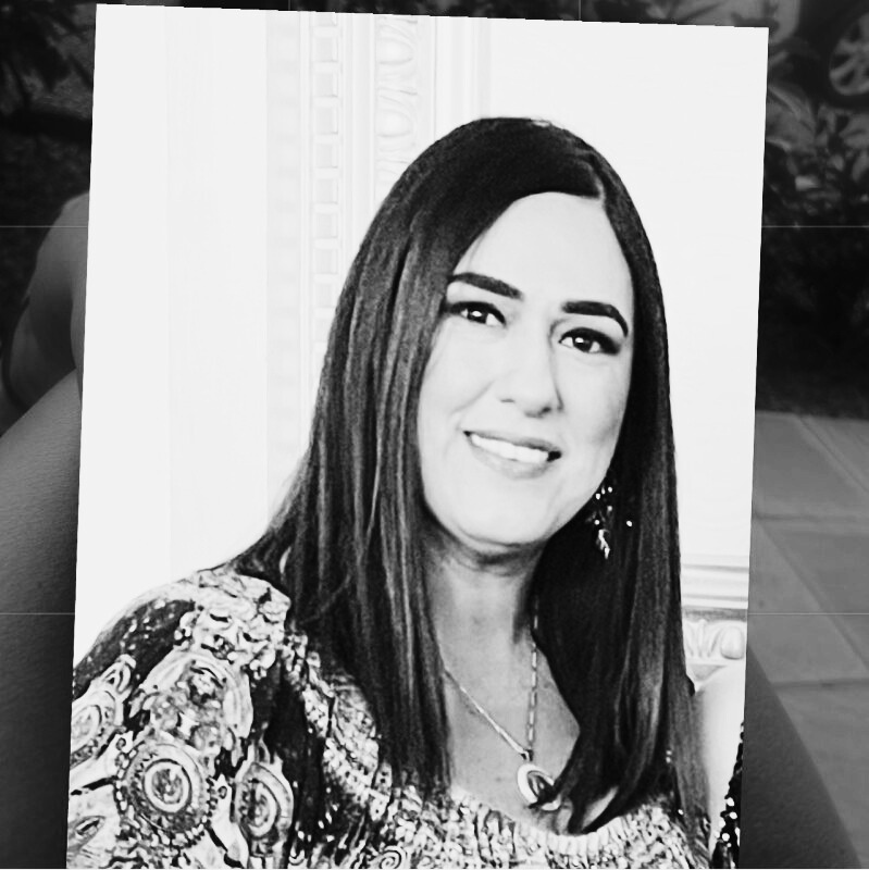 Black and white photo of a woman with long straight hair, wearing a patterned top and necklace, smiling at the camera.