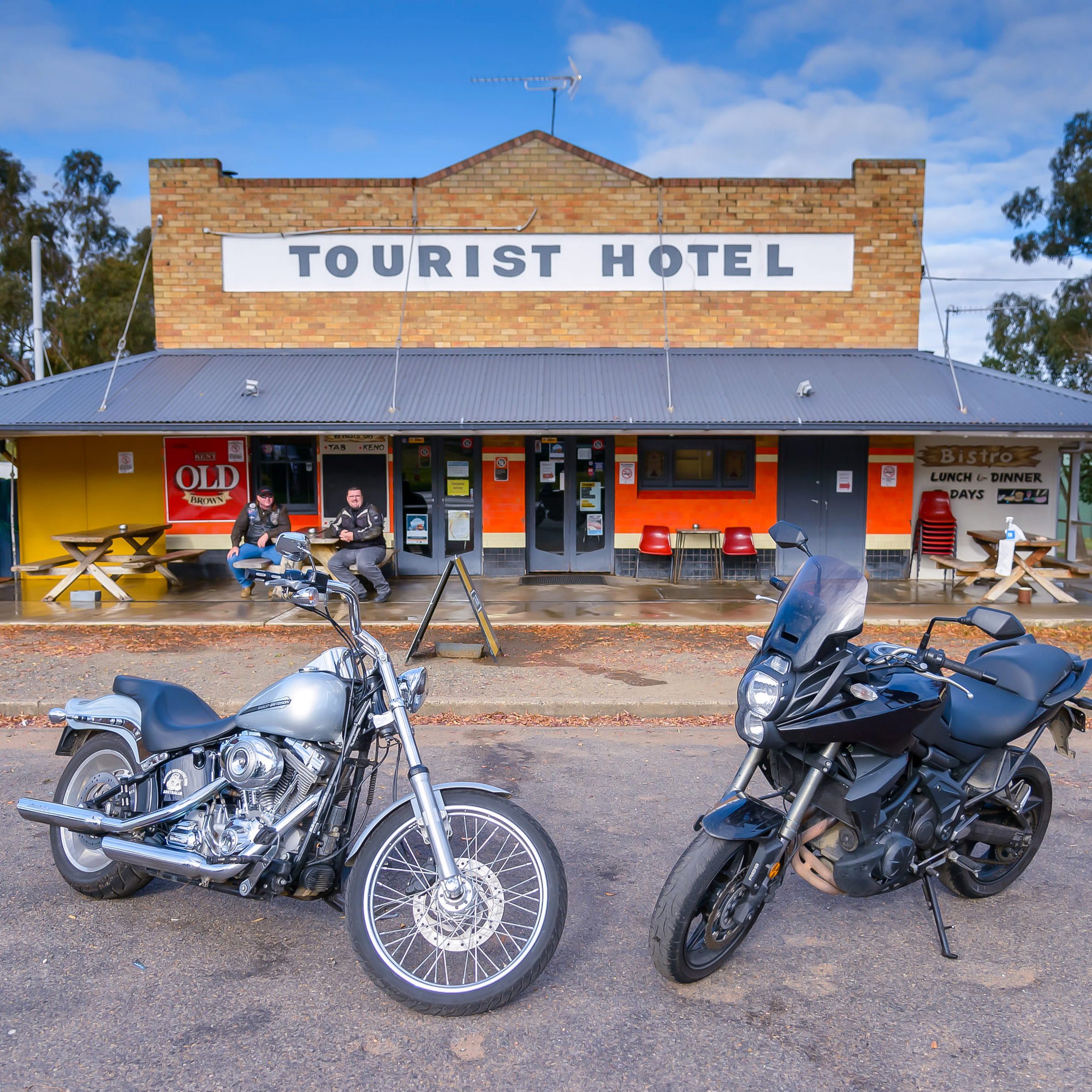 Two motorcycles parked in front of a vintage-style tourist hotel, with two people sitting on benches outside.