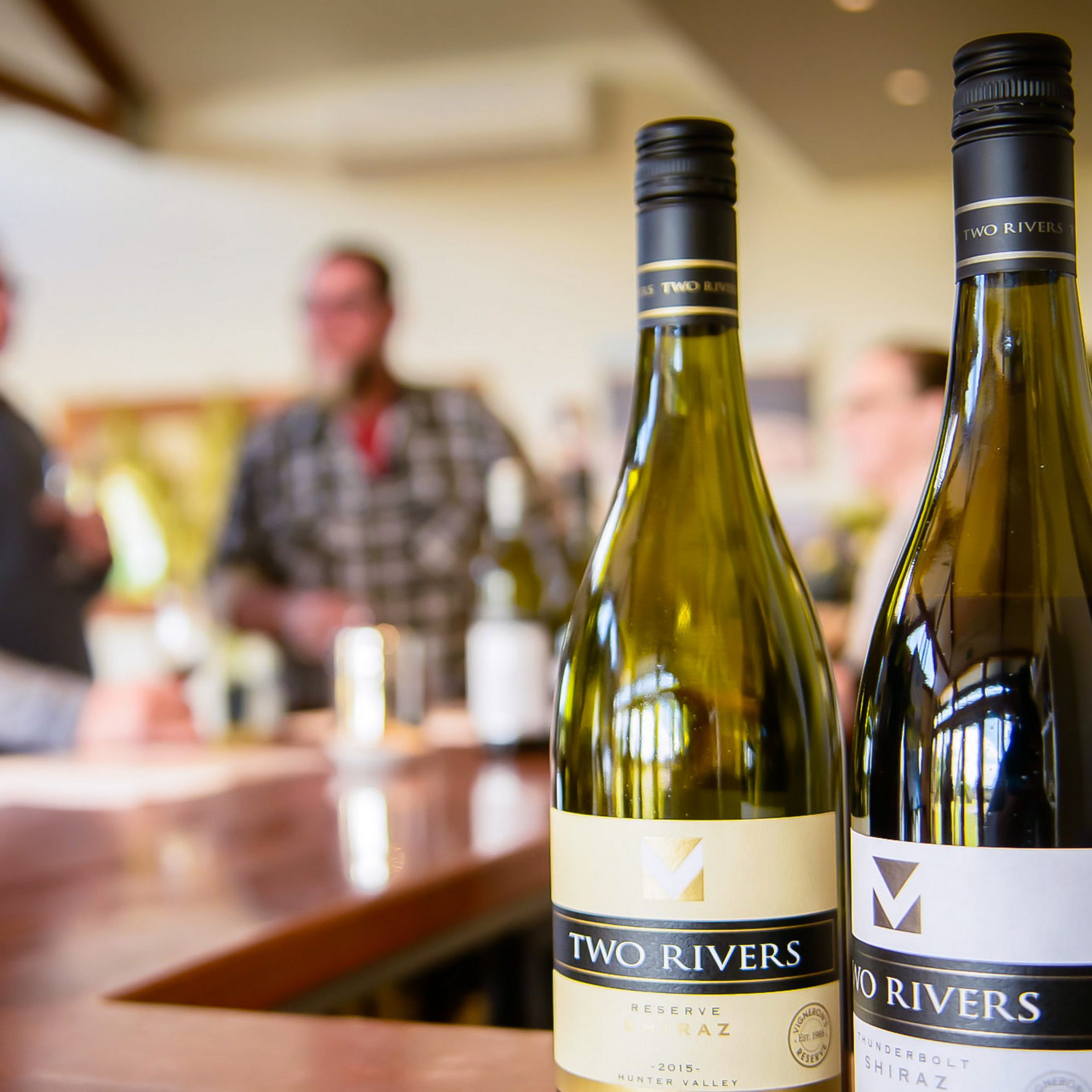 Close-up of two "Two Rivers" wine bottles on a counter with people in the background.