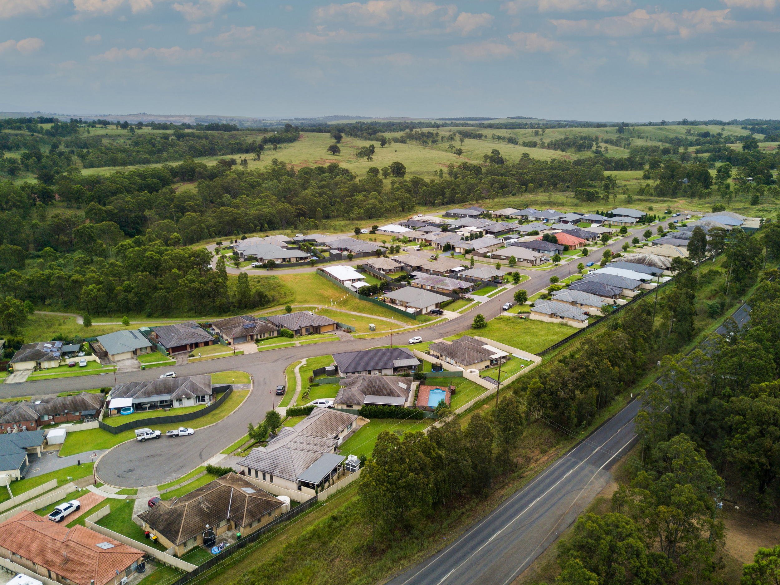 Aerial view of a suburban neighborhood with houses, roads, and surrounding green fields and forests.
