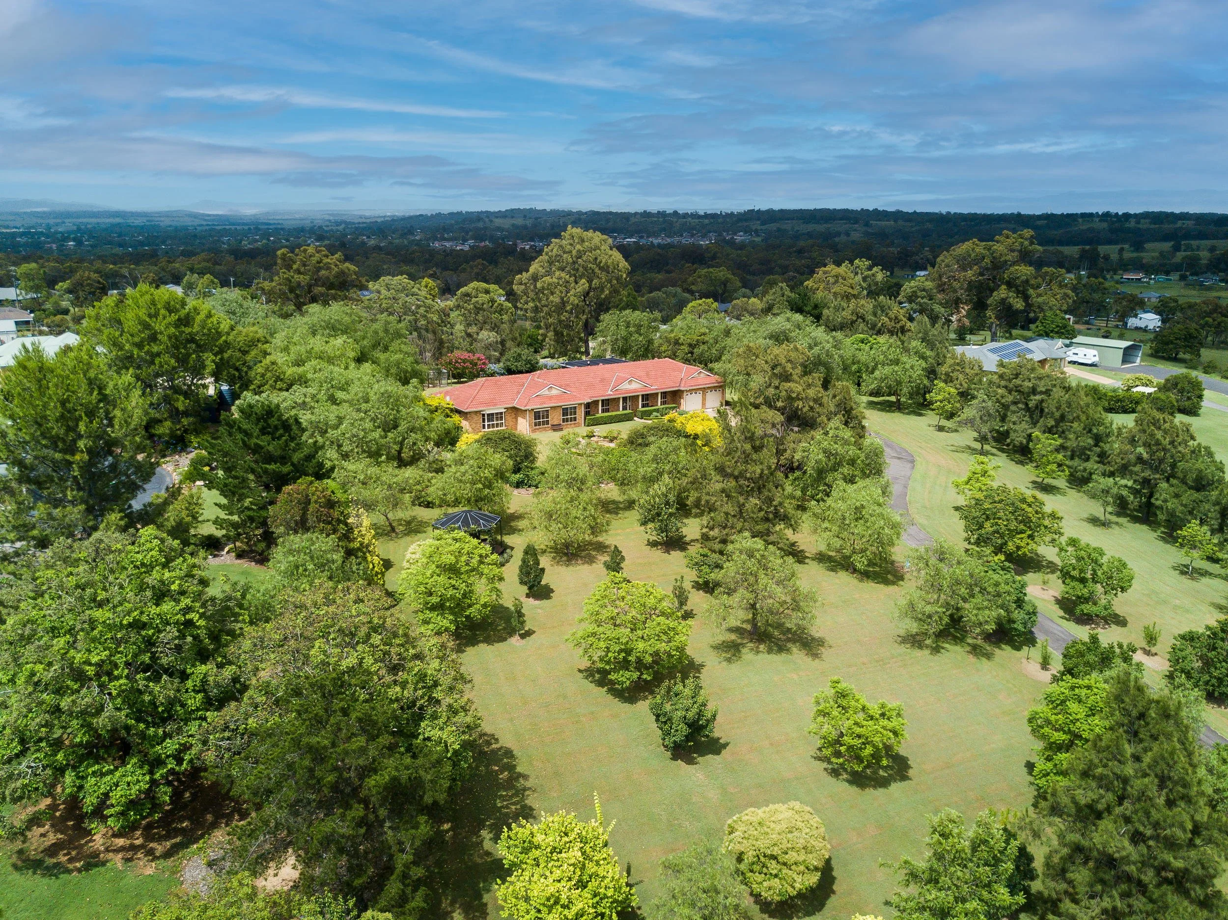 Aerial view of a house with a red roof surrounded by lush green trees and grass, set in a rural landscape.