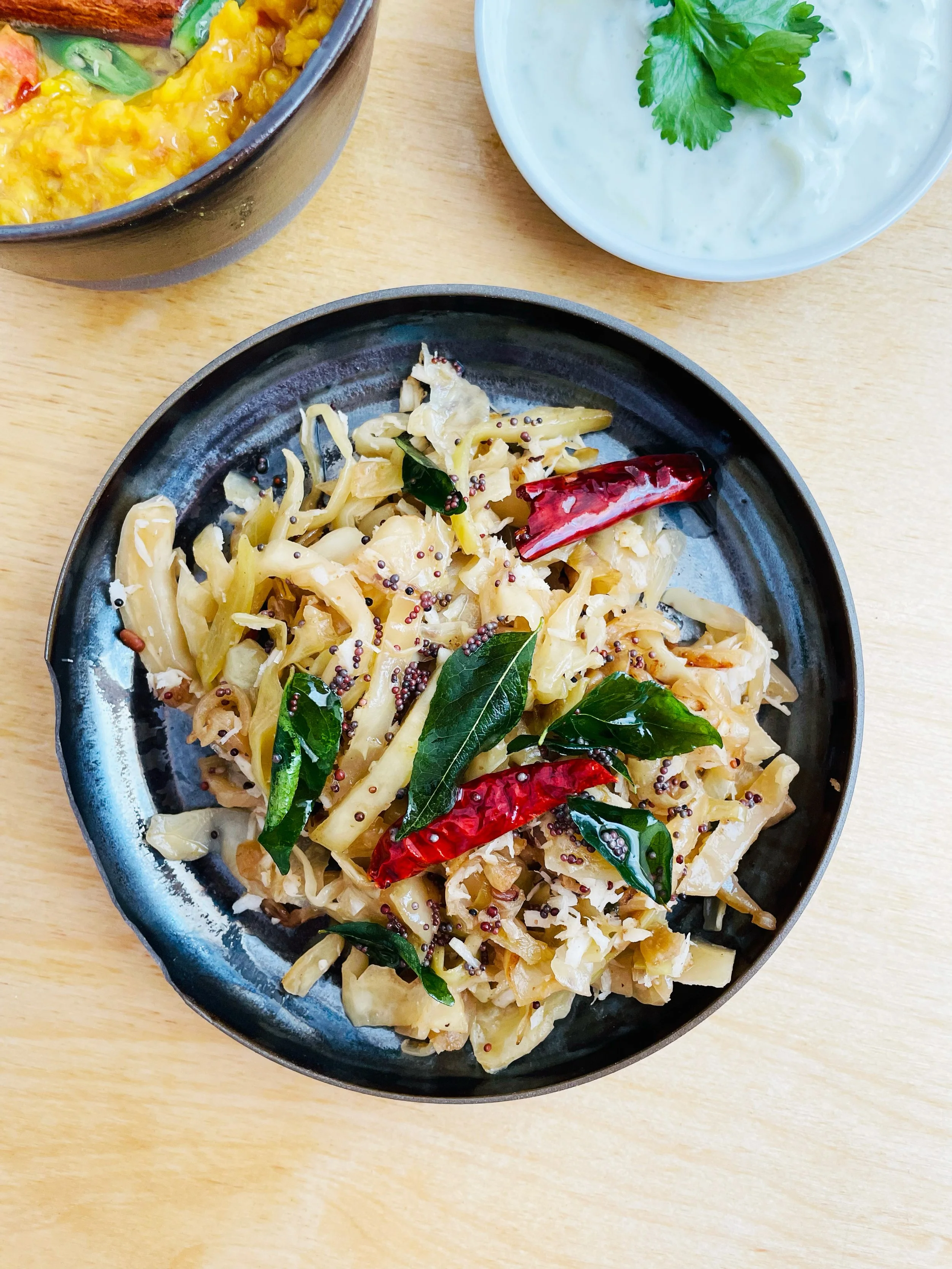 Teller mit gebratenem Weißkohl, verfeinert mit getrockneten roten Chilischoten, frischen Curryblättern und Senfsamen; im Hintergrund eine Schale mit Joghurtsoße und frischem Koriander