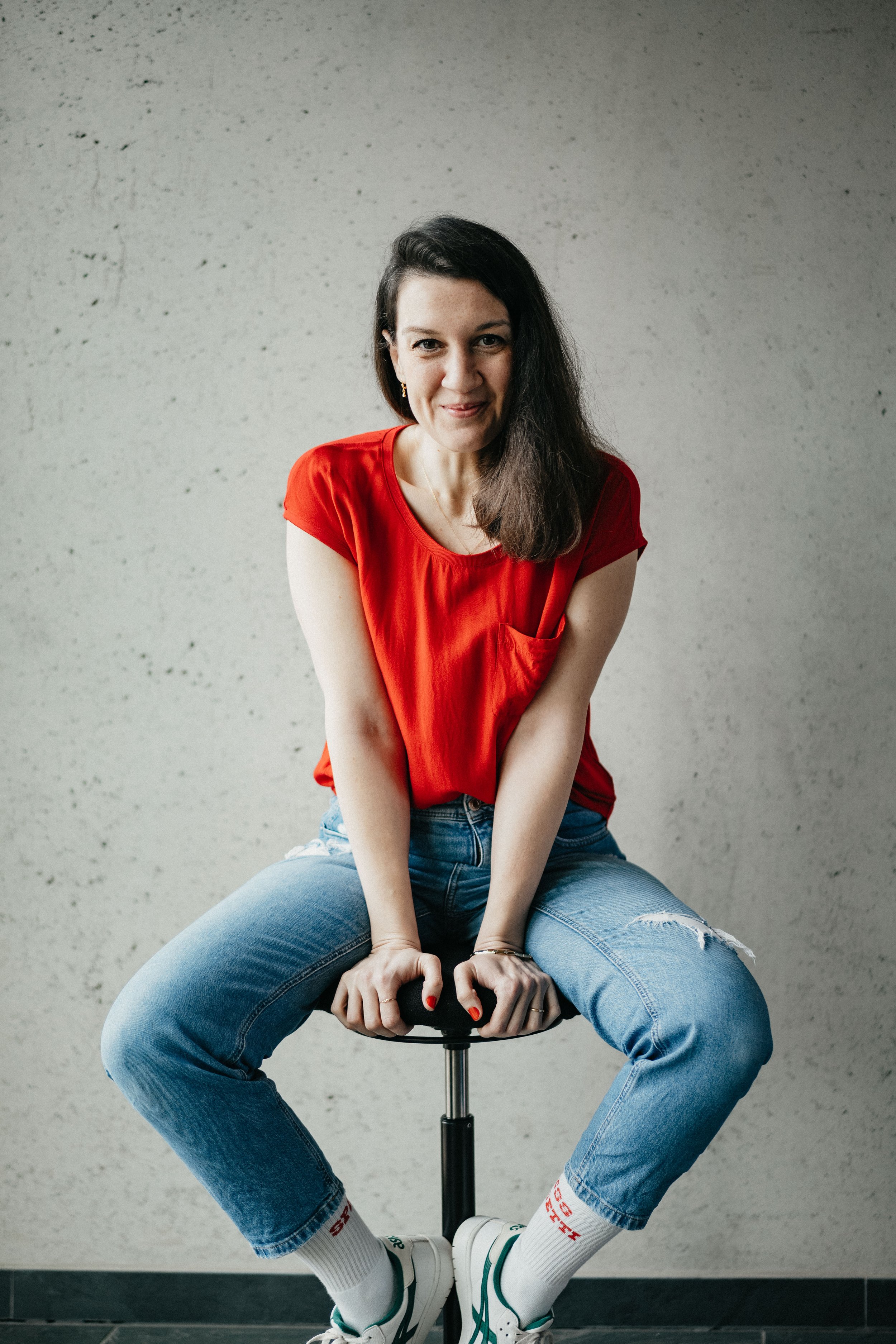 Frau in rotem T-Shirt und blauer Jeans sitzt lächelnd auf einem Hocker vor einer grauen Wand.