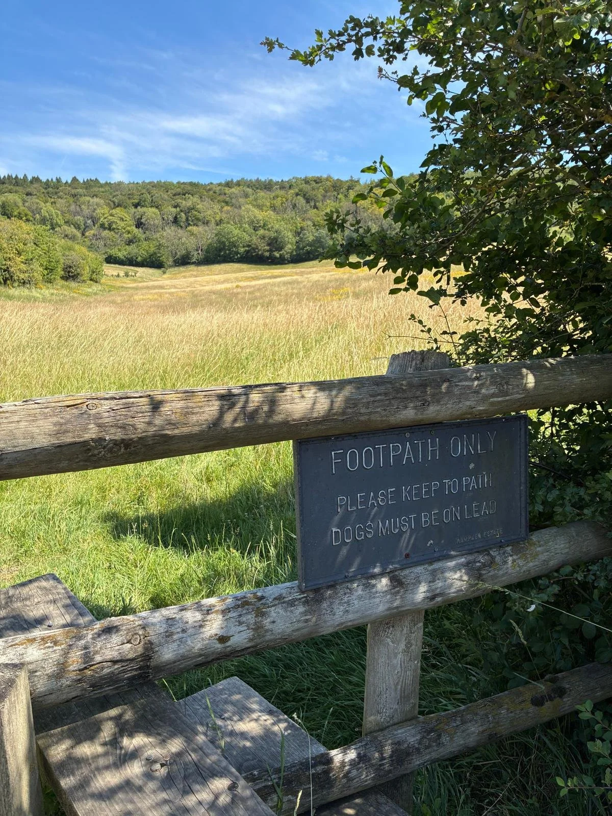 A rustic wooden fence with a sign that reads 'Footpath Only, Please Keep to Path, Dogs Must Be on Lead,' overlooking a grassy field with trees and a blue sky.