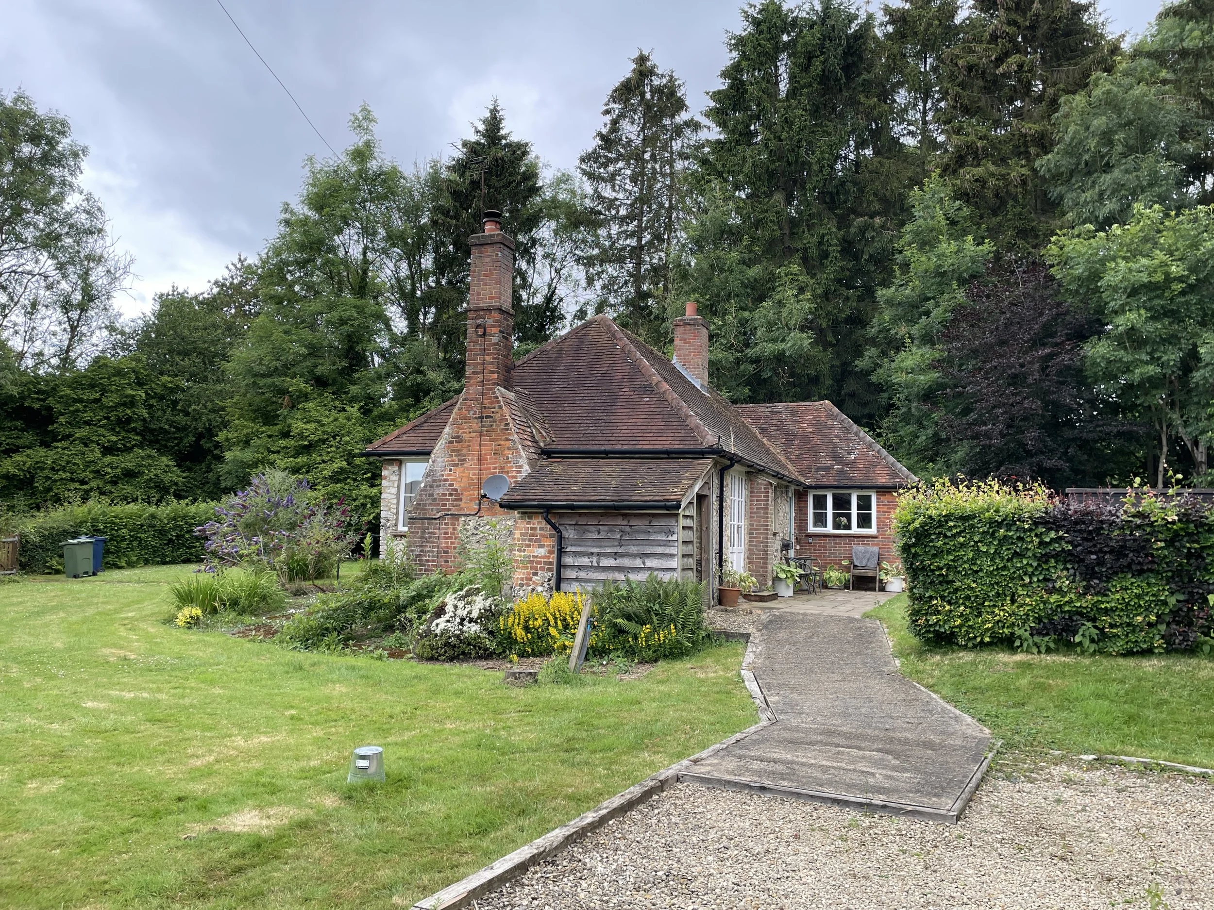 A cozy cottage with a brick and wooden exterior, surrounded by a well-maintained garden with colorful flowers and tall trees, and a gravel and concrete pathway leading to the entrance.