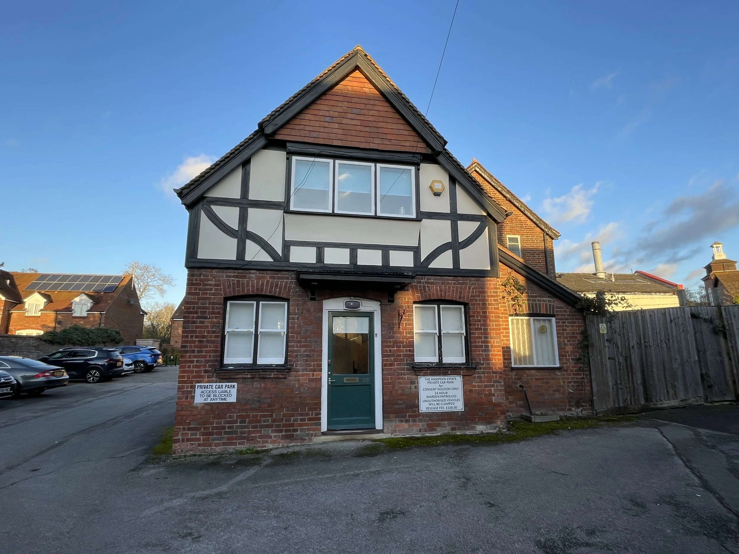 A brick house with white and black half-timbered detailing on the upper level, three white-framed windows on the lower level, and a green front door. There are cars parked to the left and signs on the house indicating it is a private car park.