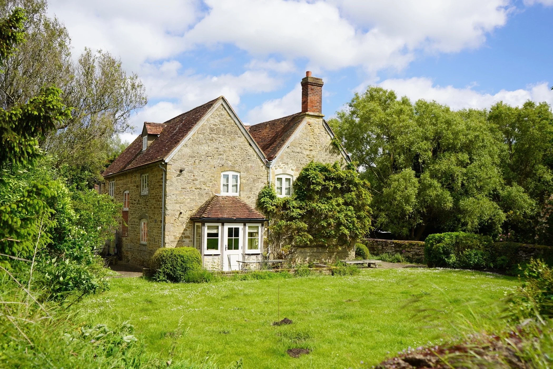 A stone house with a red-tiled roof and white window frames, surrounded by lush green trees and grass, under a partly cloudy sky.