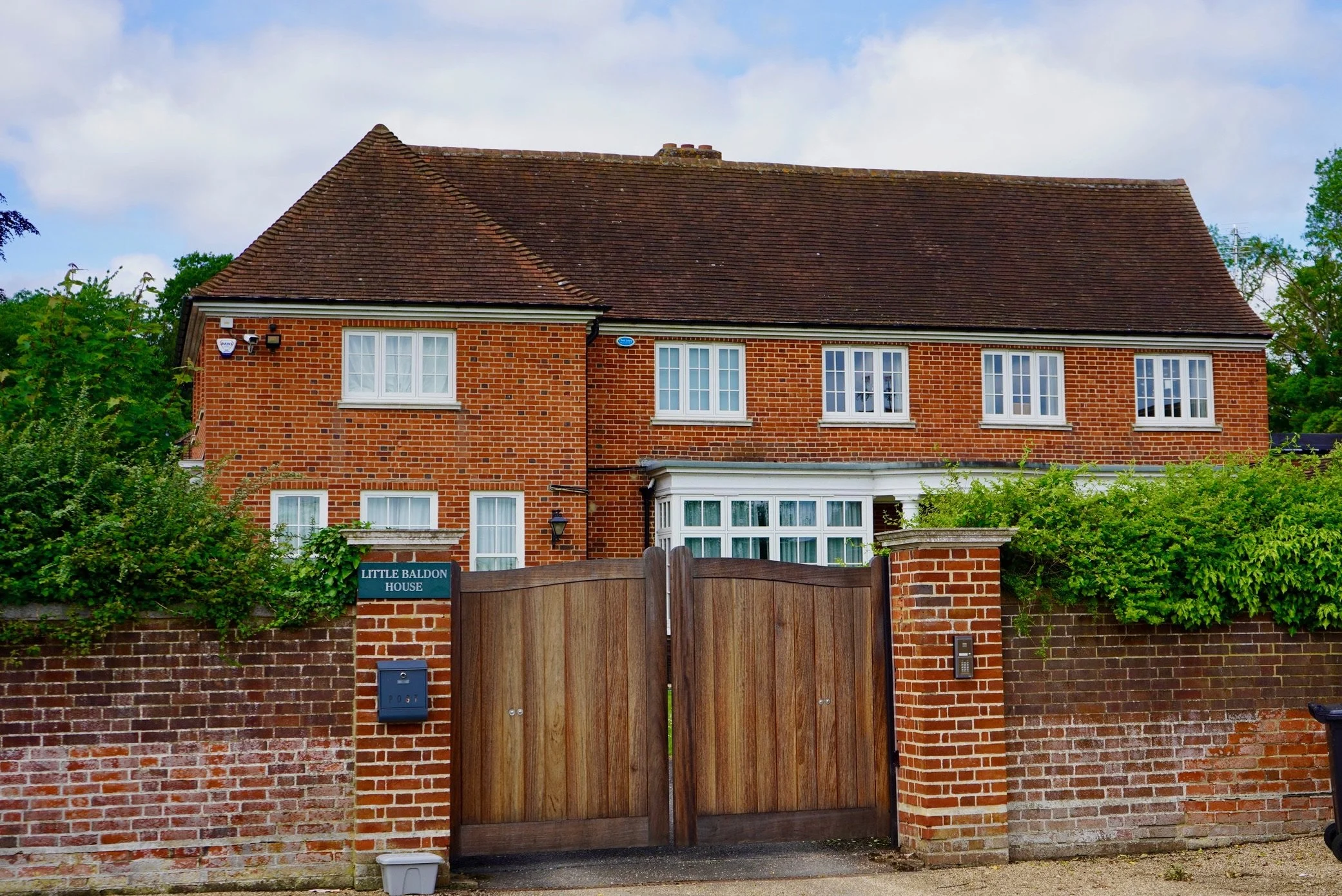 A large brick house with a tiled roof, white-framed windows, and a wooden gate in front. There is a sign that says 'Little Baldon House' and greenery around the fence.