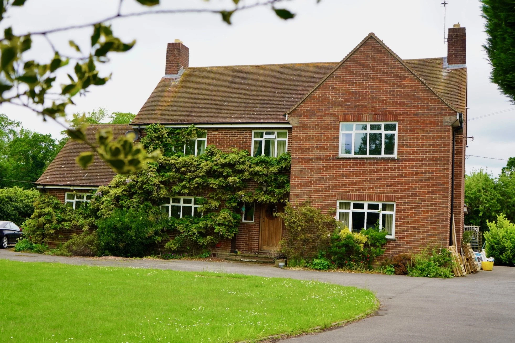 A brick house with a sloped roof, surrounded by greenery and plants, with a driveway in front.
