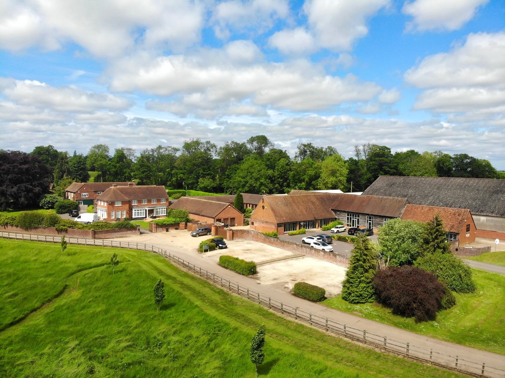 Aerial view of a rural property with brick buildings, cars parked, a gravel lot, a grassy field, and lush green trees under a partly cloudy sky.