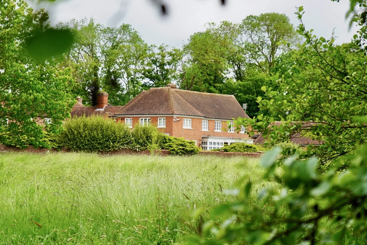 A large brick house with a sloped roof surrounded by tall trees and greenery, viewed from a grassy field with some bushes in the foreground.