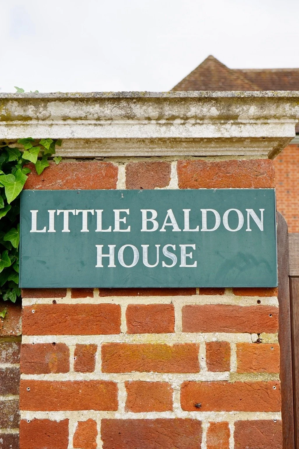 A brick wall with a green sign that reads 'Little Baldon House' and a stone cap on top, partially covered by ivy leaves.