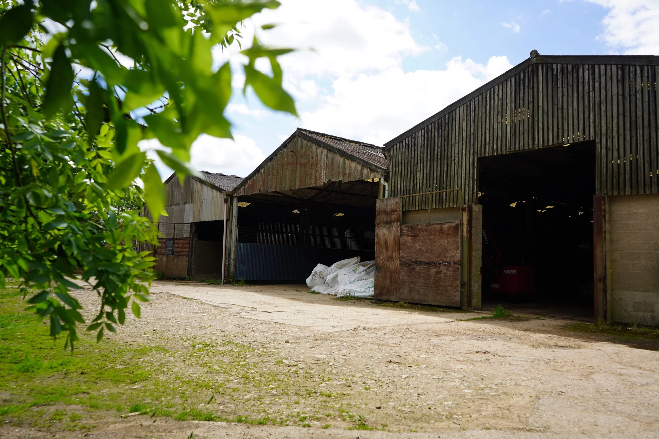 An old, partially wooden barn with several sections, some with rusted metal or weathered wood siding, open doors revealing stored items, and a dirt pathway in front with grass on the sides under a partly cloudy sky.