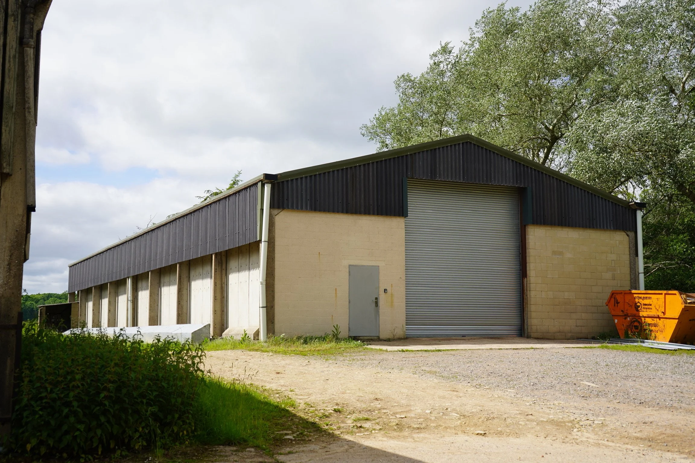 An industrial warehouse building with a metal roll-up door, a small gray door, a beige brick wall section, and an orange dumpster nearby. There are trees and a cloudy sky in the background.