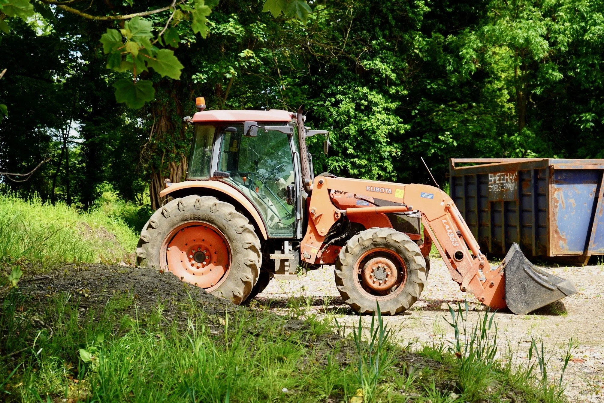Orange Kubota tractor parked outdoors next to a blue metal container, surrounded by greenery.