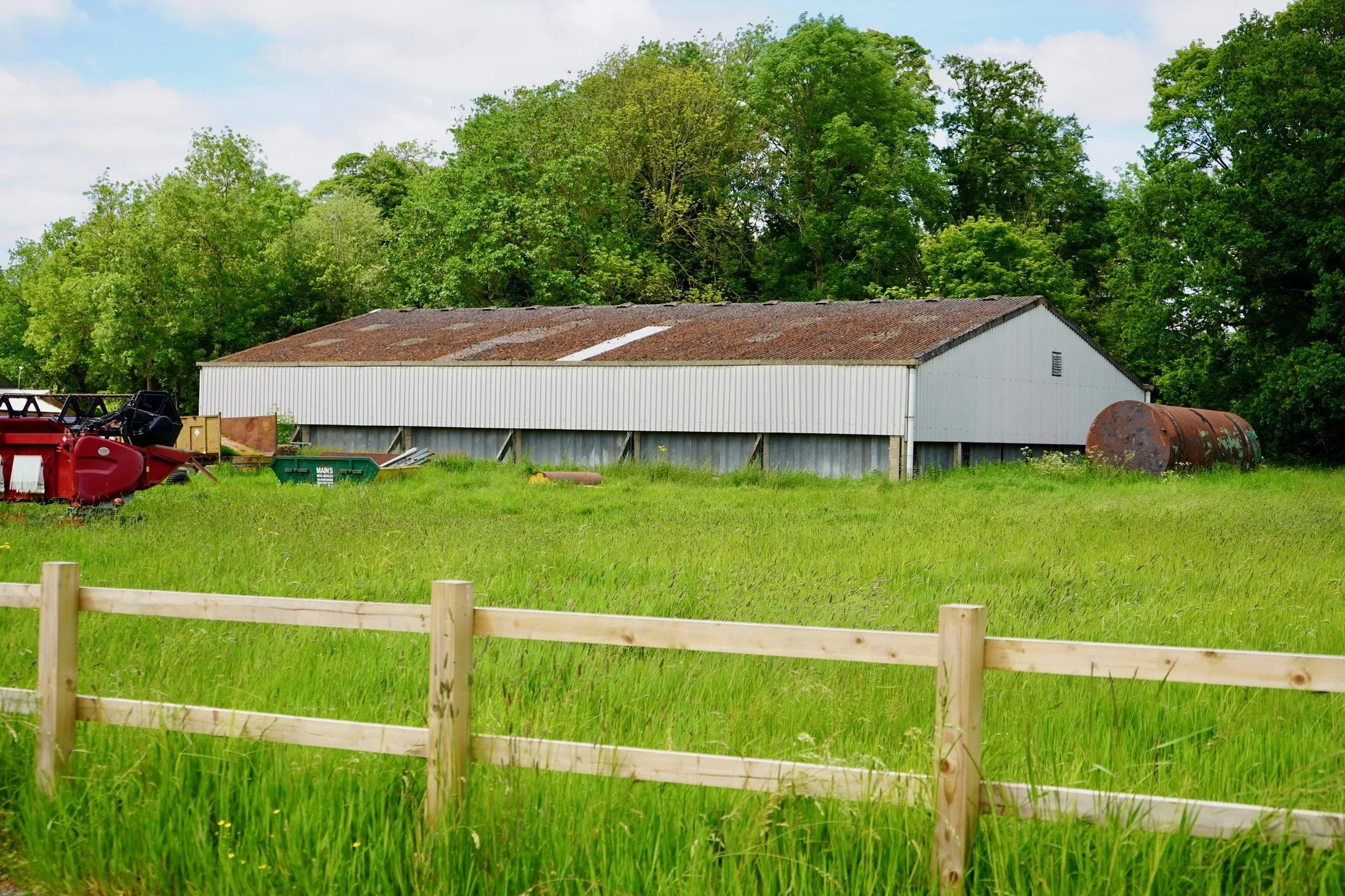 A rustic barn with a rusty roof surrounded by lush green grass and trees, with a wooden fence and machinery nearby.