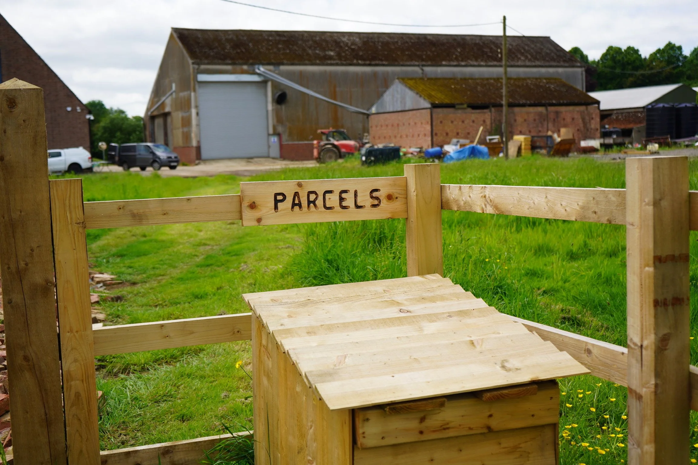 Wooden parcel delivery box with "PARCELS" sign in a rural setting with barns and vehicles in the background.