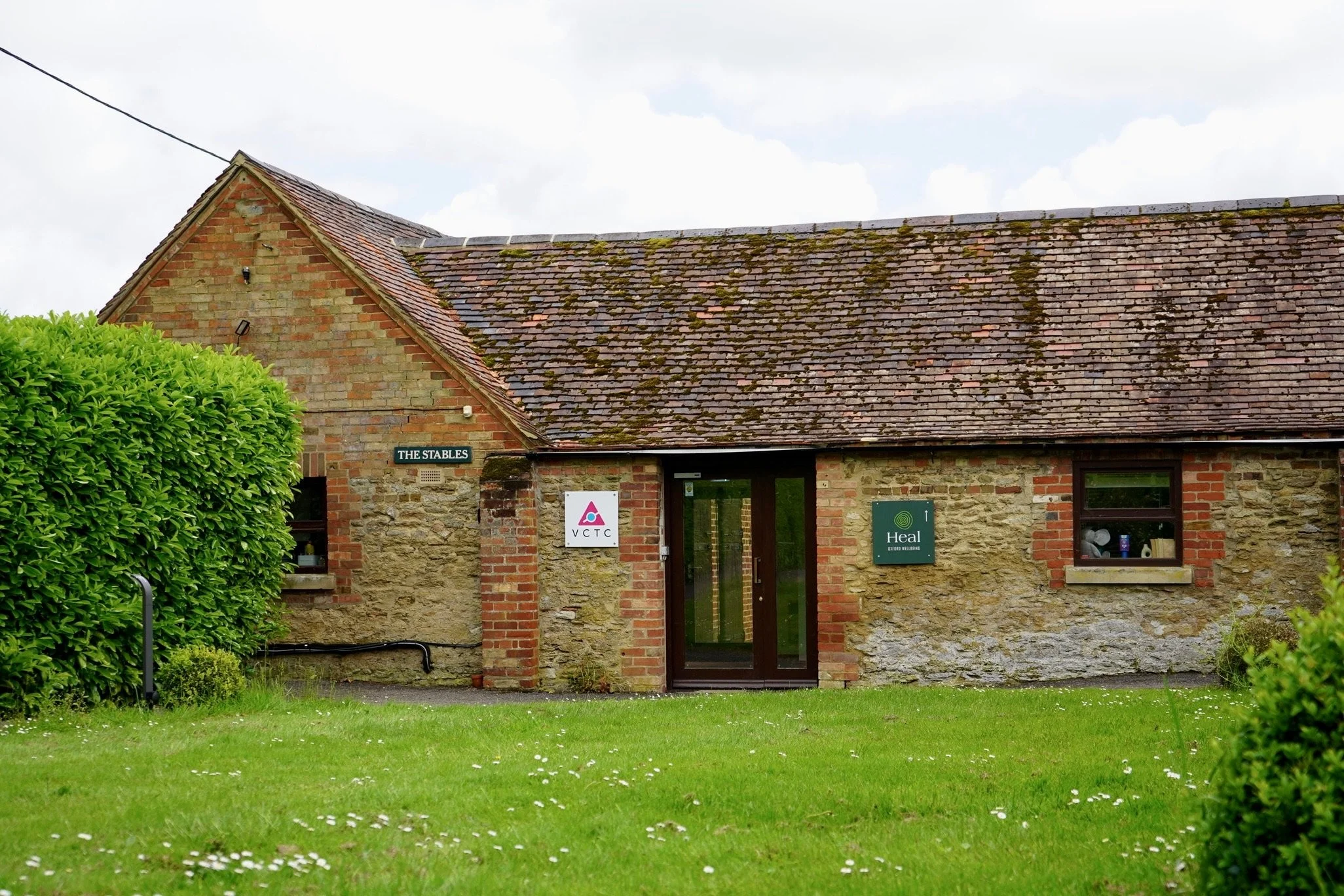 Traditional brick building labeled 'The Stables' with signs for VCTC and Heal on a grassy lawn.