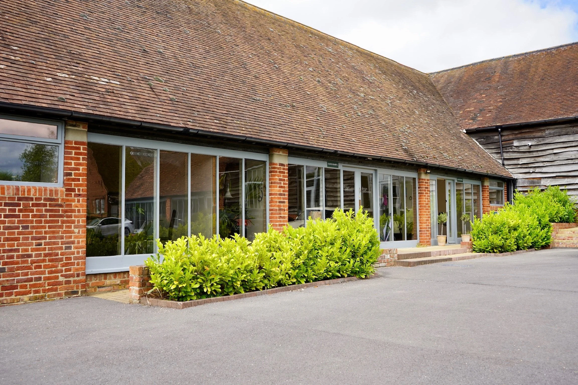 Exterior of a brick building with large glass windows, surrounded by green shrubs and a paved area.