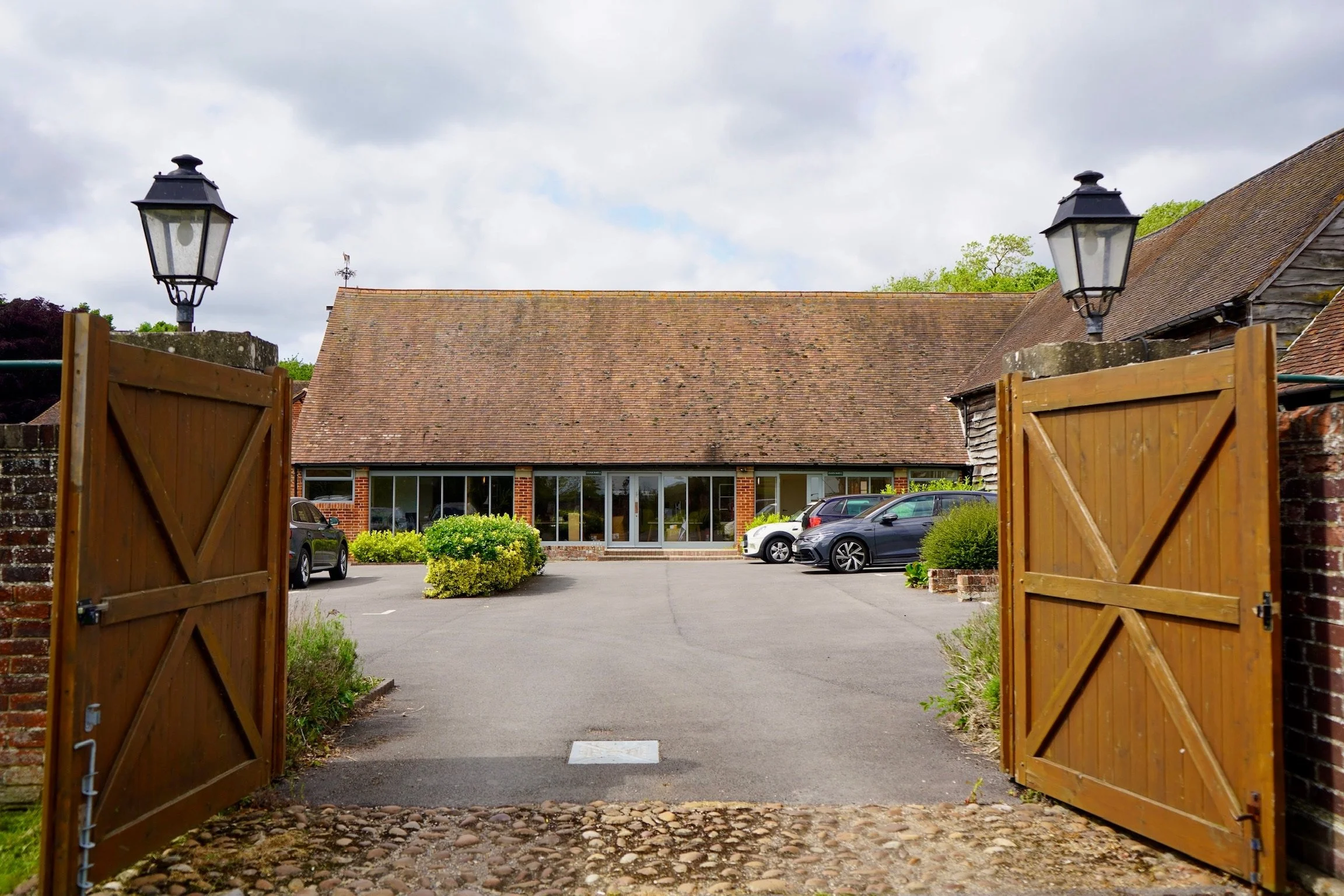 Countryside driveway leading to a brick building with a sloped roof, flanked by wooden gates and lanterns, with parked cars and greenery.