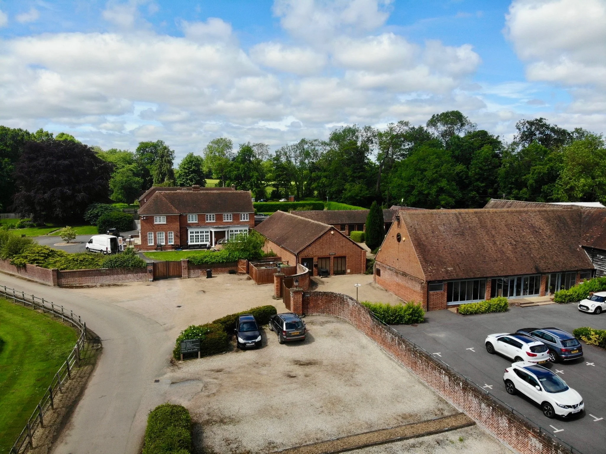 Aerial view of a countryside property with brick buildings, parked cars, and surrounding greenery on a sunny day.