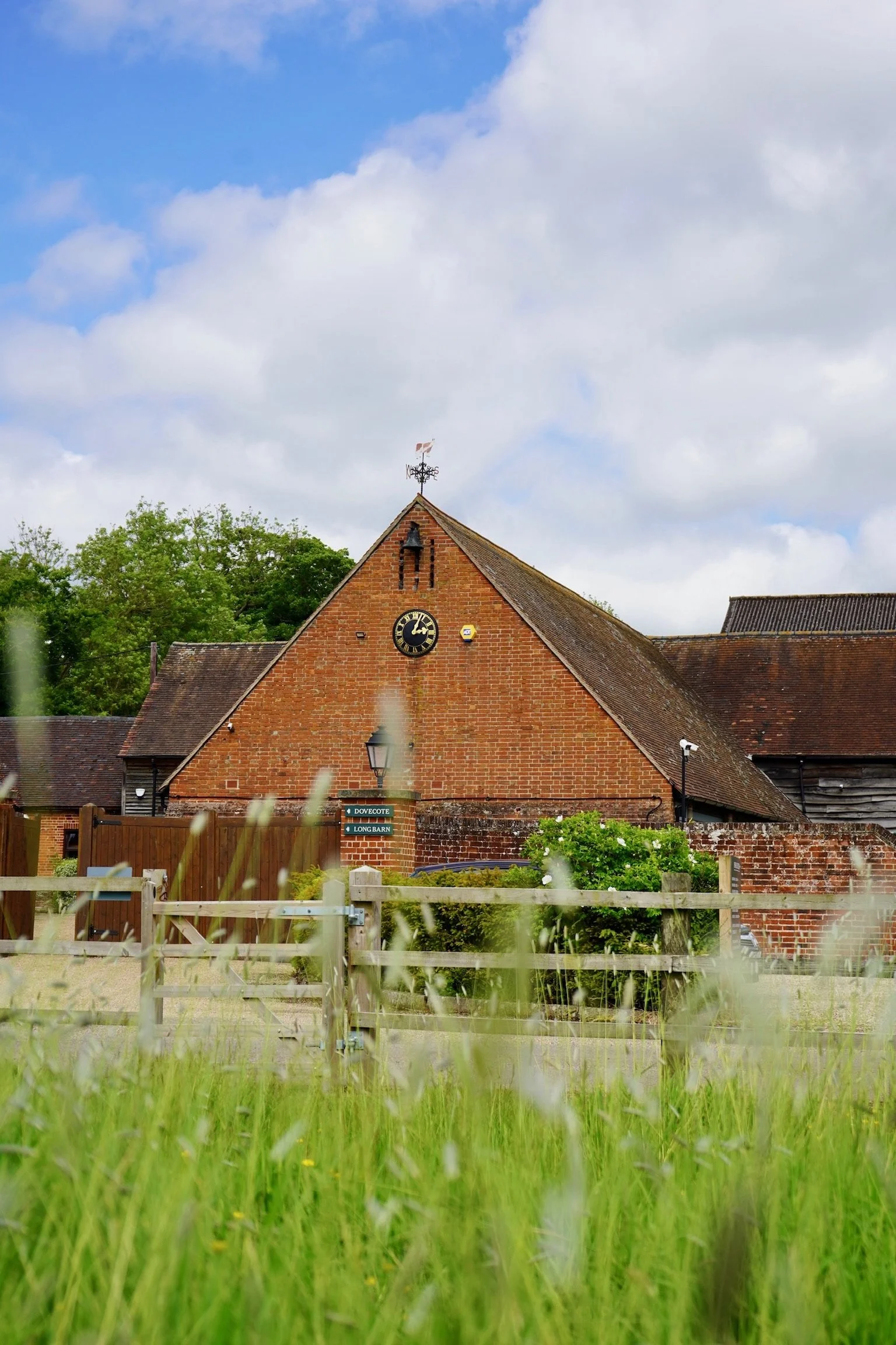 Brick building with a triangle roof and weather vane, clock on wall, surrounded by greenery and wooden fence; cloudy sky background.