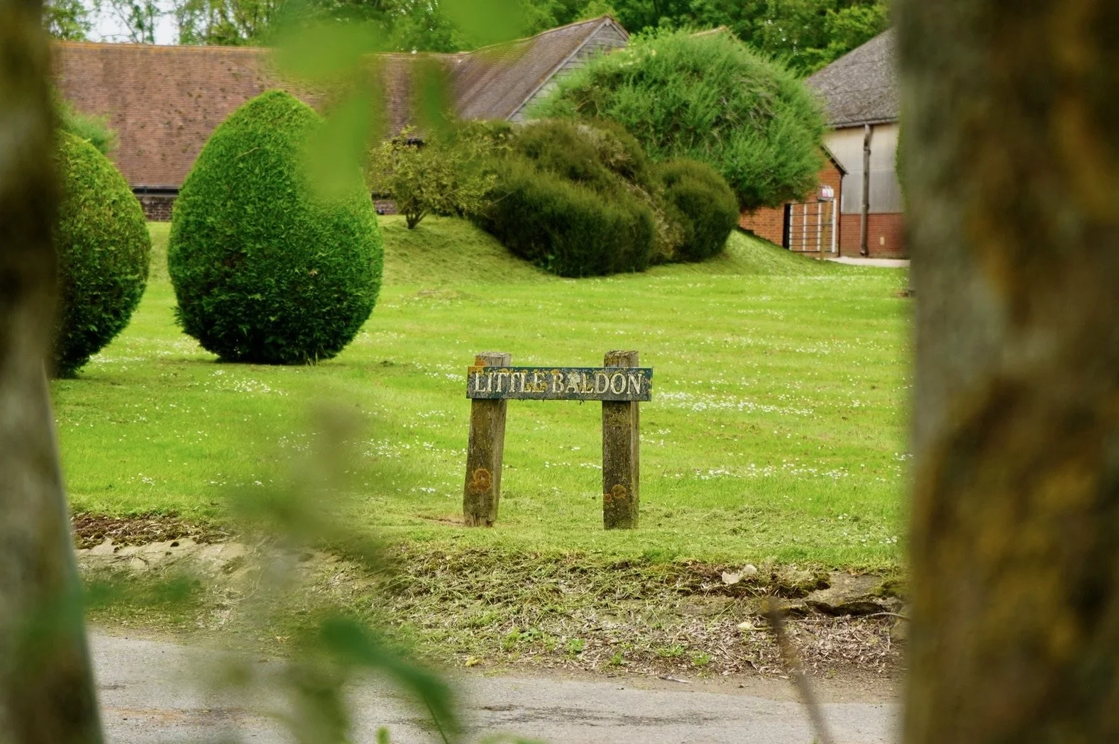 Sign reading 'Little Baldon' in a garden with trimmed bushes and a background of rural buildings.