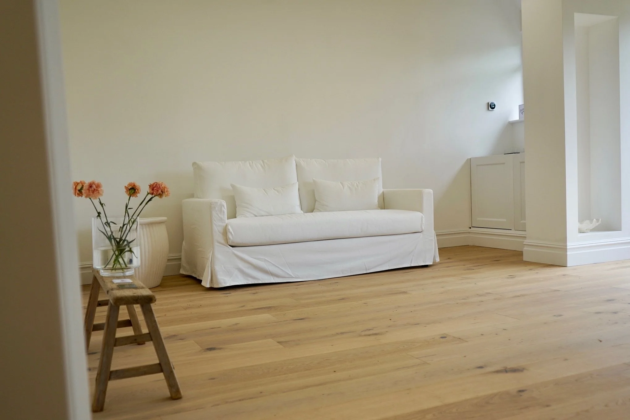 Minimalist living room with white sofa, wooden stool, flowers in vase, and hardwood floor.
