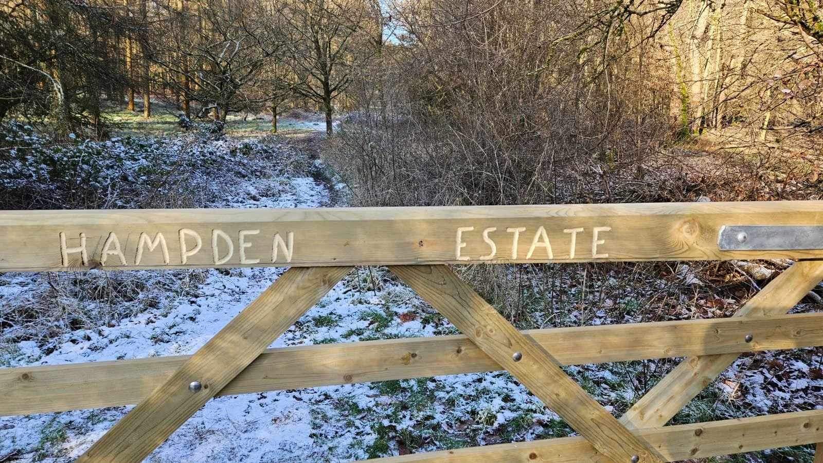 Wooden gate with 'HAMPDEN ESTATE' carved into it, set in a snowy forested area with trees and bushes in the background.