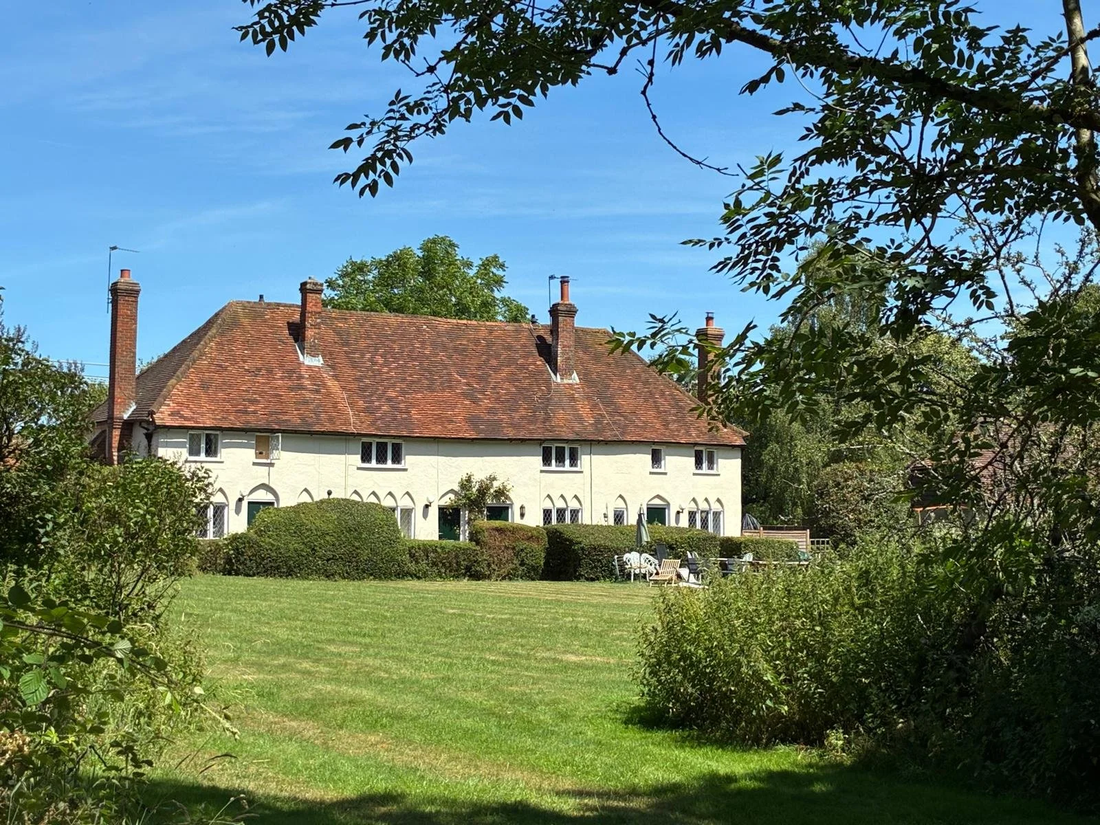 A white house with a red-tiled roof and chimneys, surrounded by greenery and a well-maintained lawn, under a clear blue sky.
