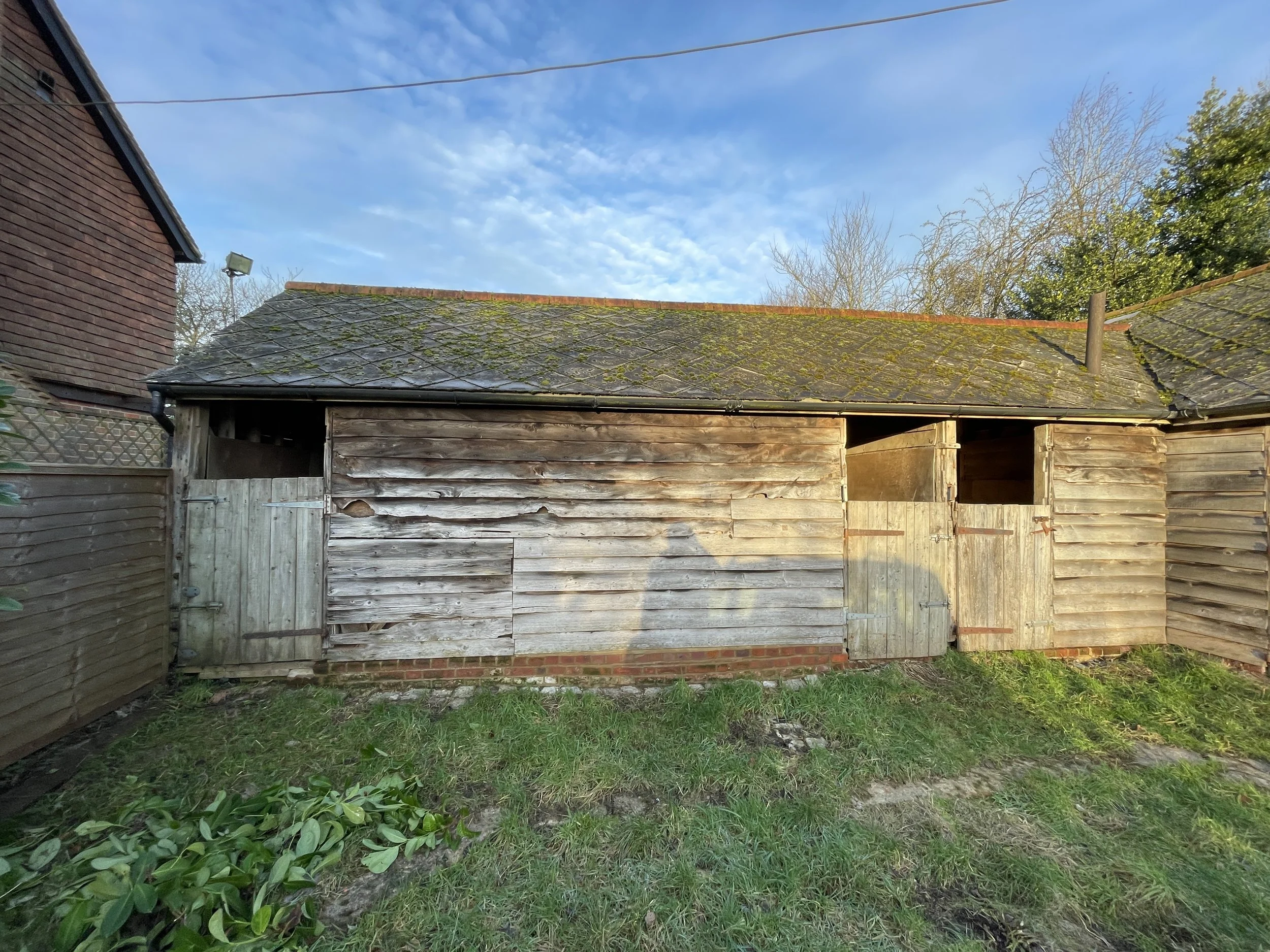 An old wooden shed with weathered, grayish planks, some of which are warped and damaged. The shed has a moss-covered, slightly sloped shingled roof and two small entrances with doors, one of which is slightly open. Surrounding the shed are a grassy y