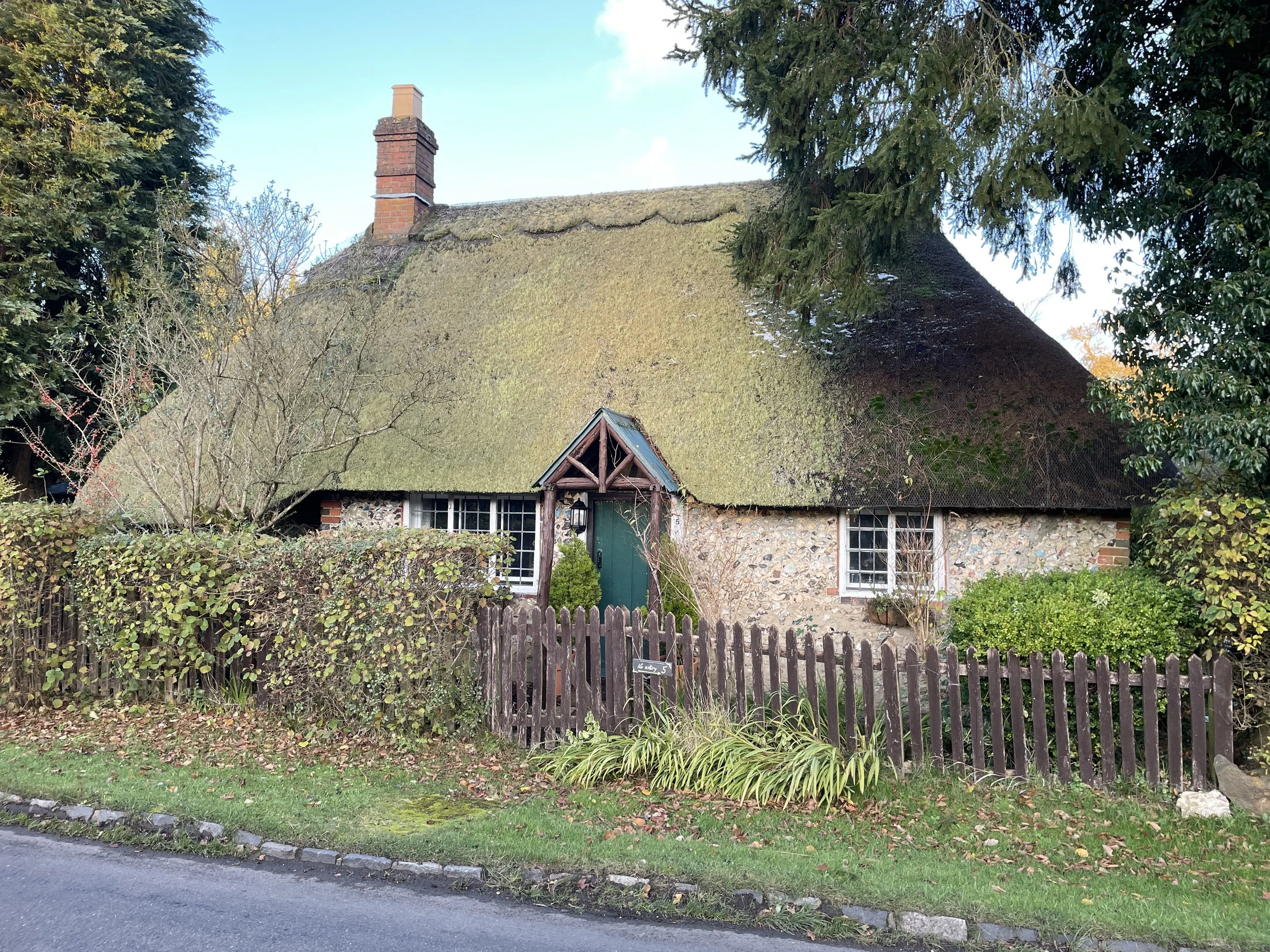 A cottage with a thatched roof, stone walls, and white-framed windows, surrounded by a garden and a wooden fence, in a suburban neighborhood.
