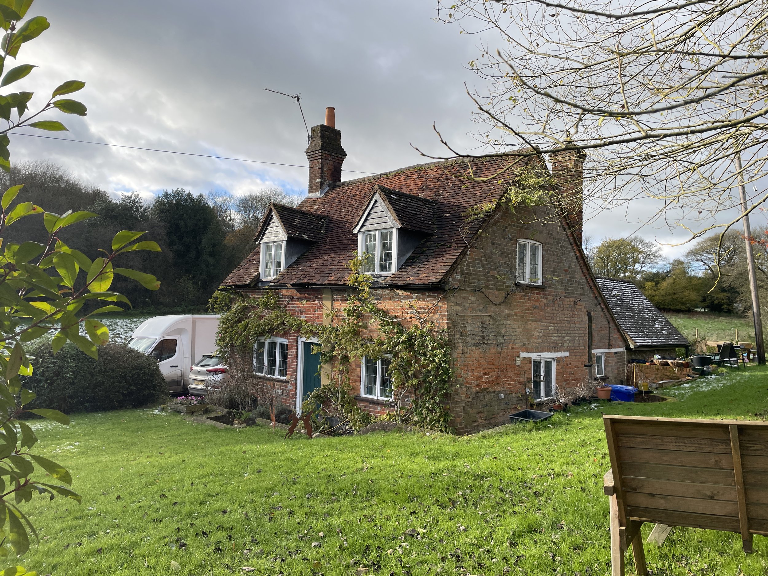 A rustic brick house with a sloped roof, dormer windows, and a chimney, surrounded by a green lawn and trees in a countryside setting.