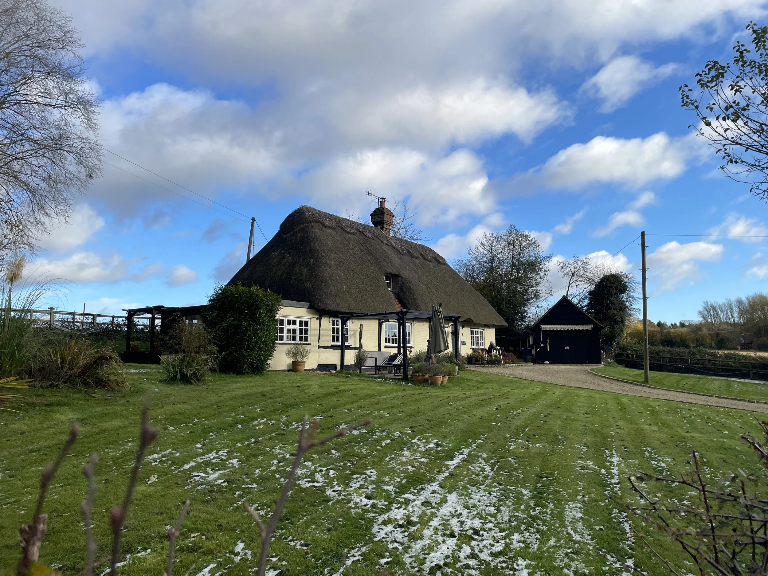 A quaint countryside house with a thatched roof, white walls, and multiple windows. The house is surrounded by a green lawn with patches of snow, trees, and shrubs. There is a gravel driveway leading to a small black shed and a wooden fence in the background under a partly cloudy sky.
