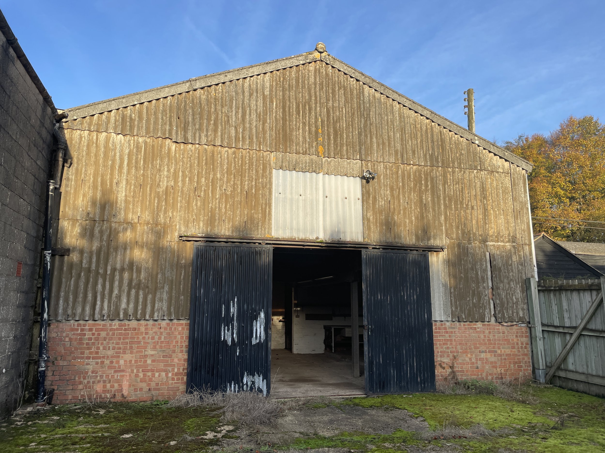 An old, weathered barn with corrugated metal siding, partially open black doors, brick foundation, and surrounded by mossy ground under a blue sky with some trees in the background.
